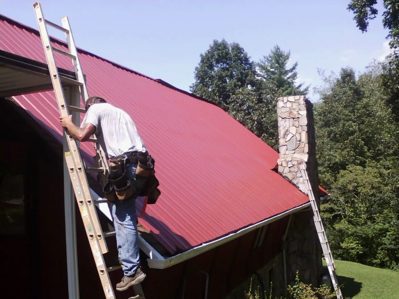 Roofer on a ladder beside a red metal roof and stone chimney — Soffit & Fascia Repair by Roofing Brothers in Bluffton